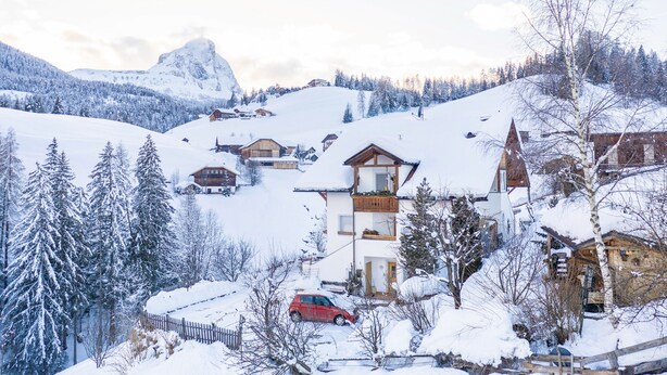 A snow-covered mountain village with wooden houses, a red car, and a prominent mountain peak in the background.