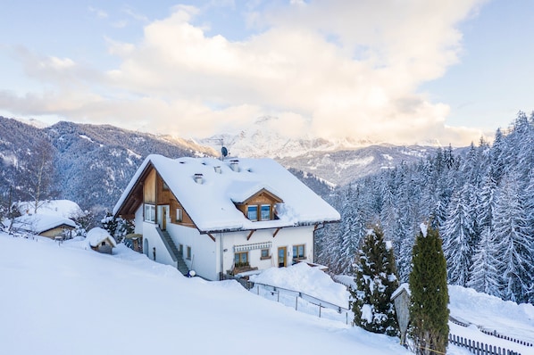 A snow-covered mountain house with a wooden exterior, a balcony, and a view of snow-covered trees and mountains.