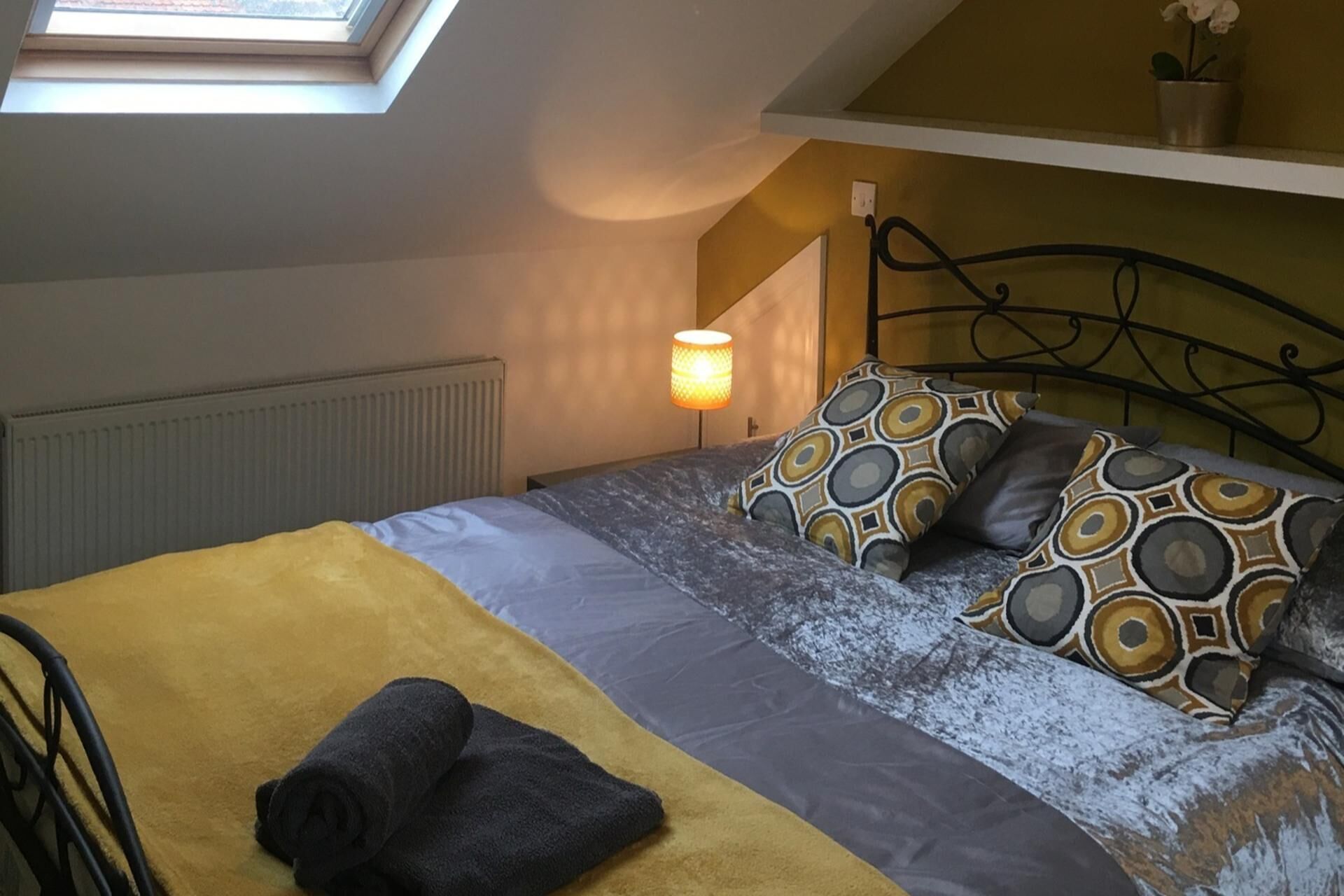Un dormitorio con tragaluces, una cama con almohadas estampadas, una lámpara de noche y un radiador.