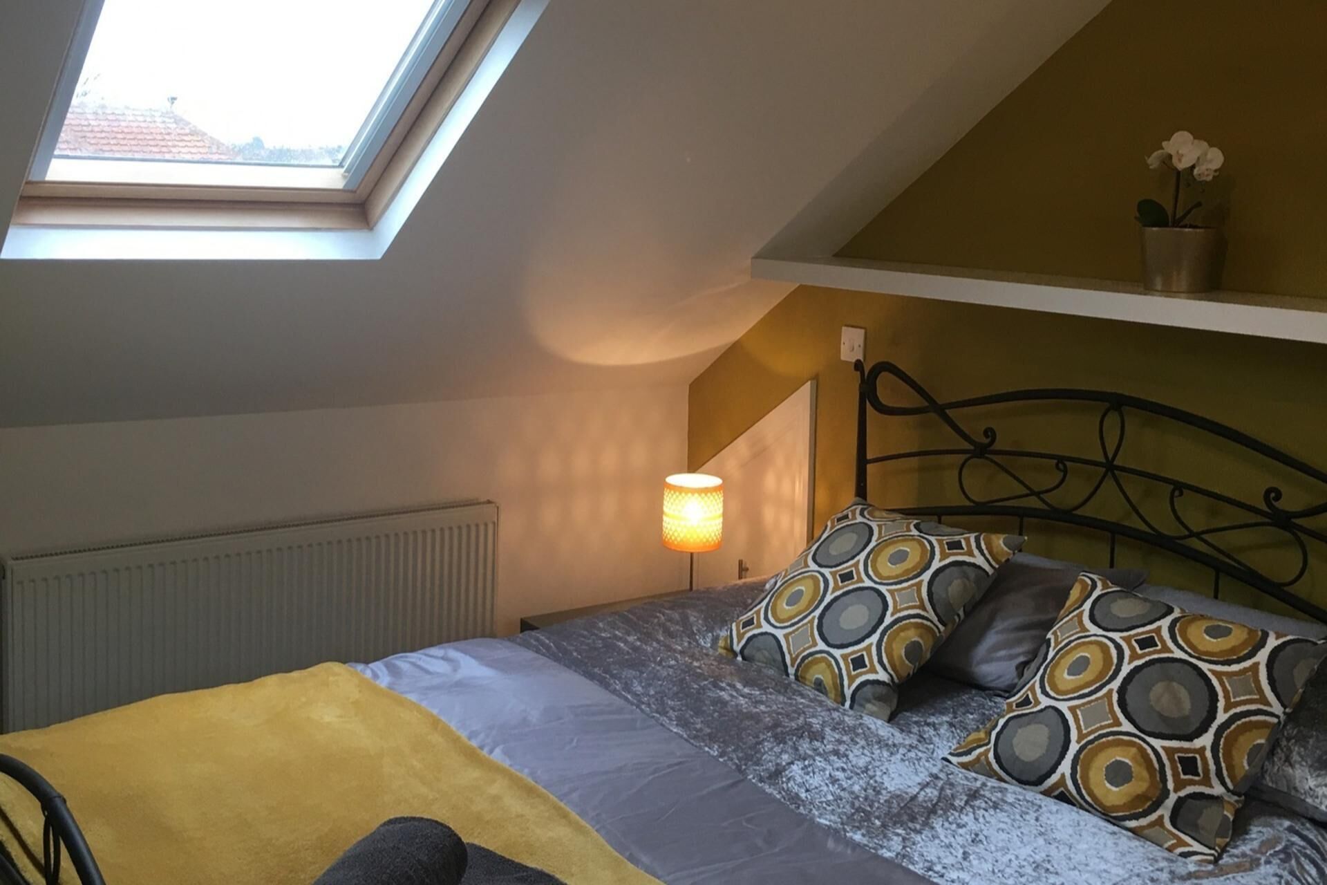 A bedroom with a skylight, a bed with patterned pillows, a bedside lamp, and a radiator.