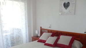 A bed with a white quilt and red pillows, a wooden headboard, a bedside table with a lamp, and a wall decoration. - Beautiful apartment facing the sea (Mont-roig del Camp)
