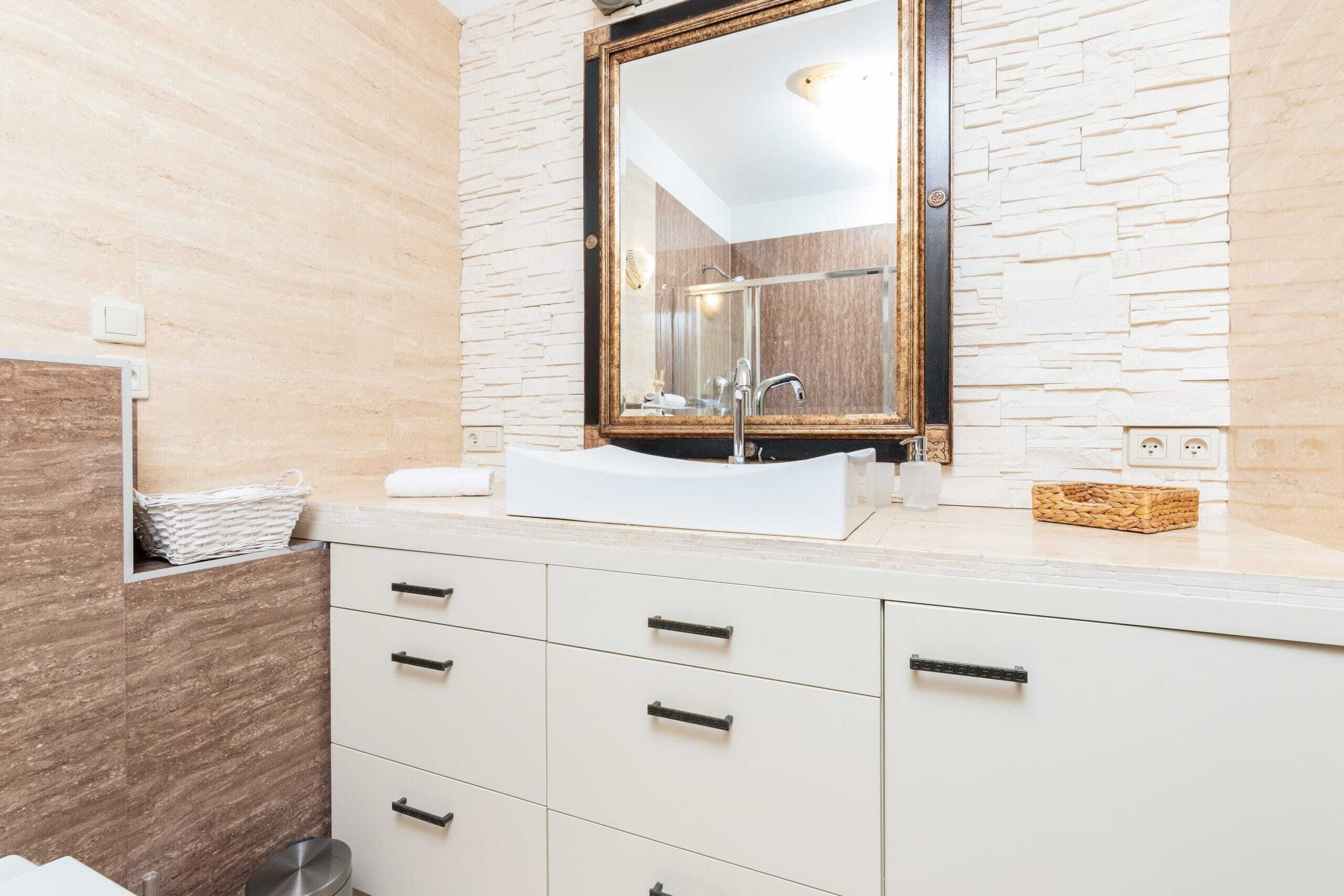 Un bagno moderno con un lavandino bianco, uno specchio grande e una parete dalla texture che ricorda la pietra.
