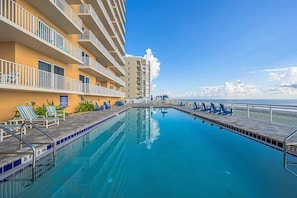 A swimming pool with lounge chairs and a deck, surrounded by a building with balconies. - 902 Sanibel - Oceanfront Panoramic Views (Daytona Beach Shores)