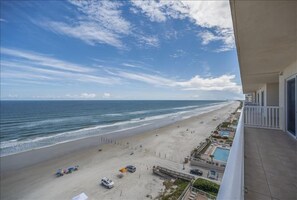 A beach view from a balcony overlooking a sandy beach with a pool and a building with a white railing. - Sanibel Condo #902 3/3 Oceanfront Panoramic Views (Daytona Beach Shores)