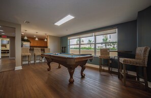 A pool table in a room with a view of palm trees through large windows, wicker chairs, and a kitchen area in the background.