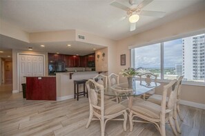 A spacious living area with a glass-top dining table surrounded by wicker chairs, a kitchen with wooden cabinets, and a large window offering a city view.
