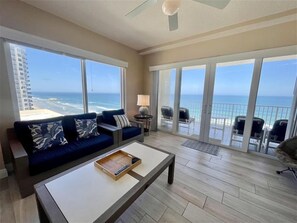 A spacious living room with a large window overlooking the ocean, a sofa with patterned cushions, a coffee table, and a balcony with chairs.