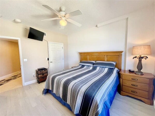 A bedroom with a bed, a nightstand, a lamp, a ceiling fan, and a TV mounted on the wall.