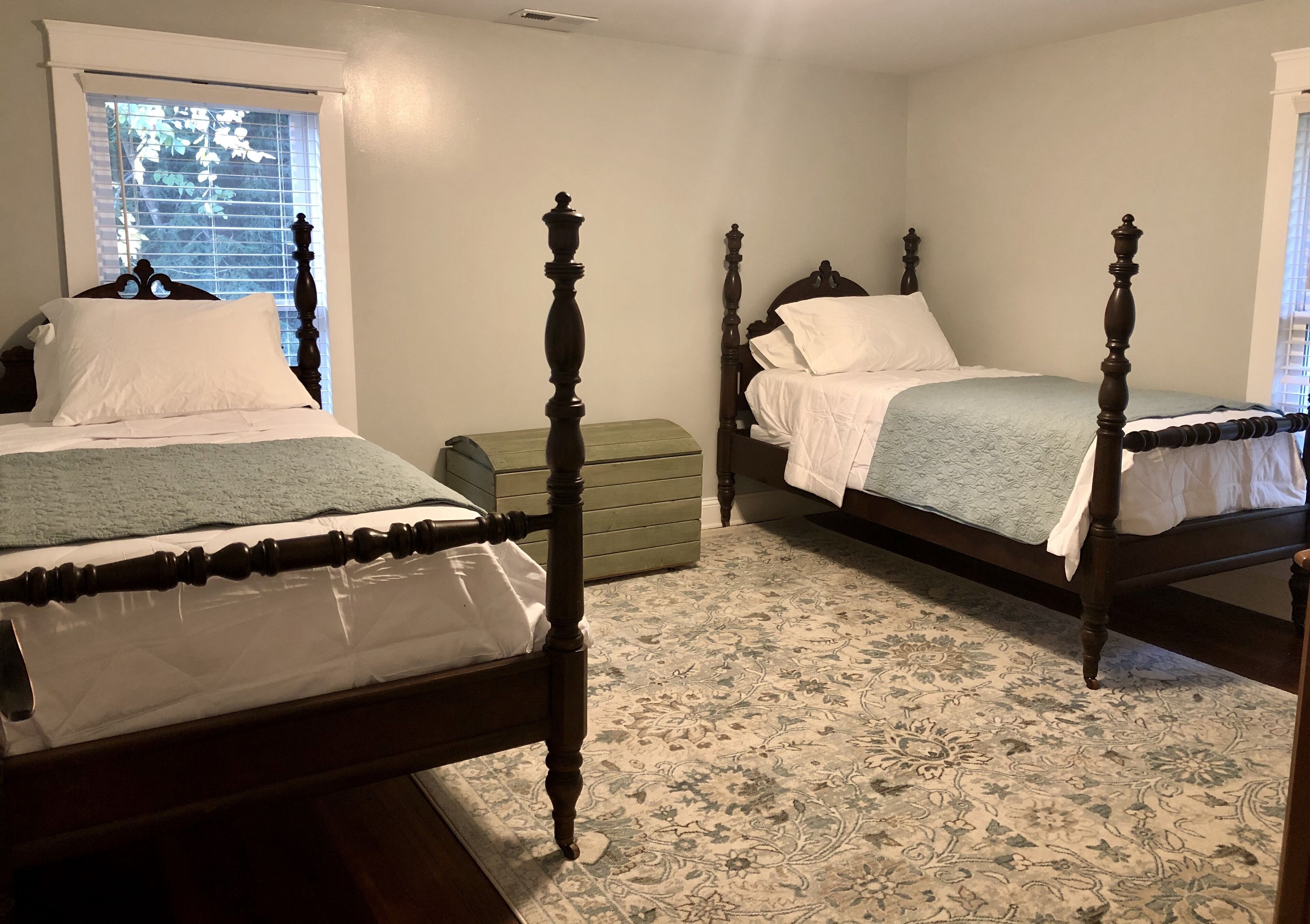 Two wooden beds with ornate posts, a green chest of drawers, and a patterned carpet in a bedroom with a window.
