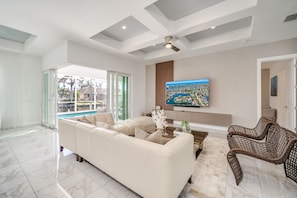 A spacious living room with a large white sofa, a coffee table, and a wicker chair. A flat-screen TV is mounted on the wall, and there is a sliding glass door leading to a pool area.