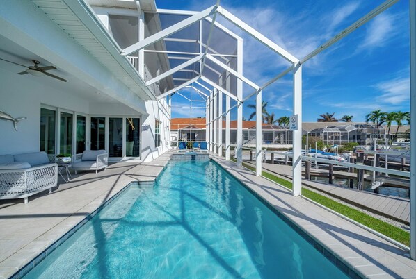 A swimming pool with lounge chairs and a covered area, surrounded by a white structure and palm trees.