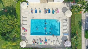 Vista aérea de una zona de piscina con múltiples sillones de descanso y sombrillas, rodeada de vegetación y un edificio al fondo.