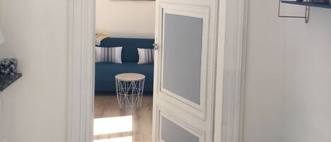 A hallway with a white door leading to a room with a blue sofa and a painting on the wall.