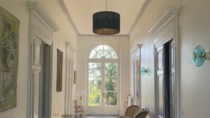 Ein geräumiger Flur mit hoher Decke, einem großen Rundbogenfenster und einer hängenden Leuchte. Es sind Korbstühle und ein Tisch mit einem Bilderrahmen zu sehen.