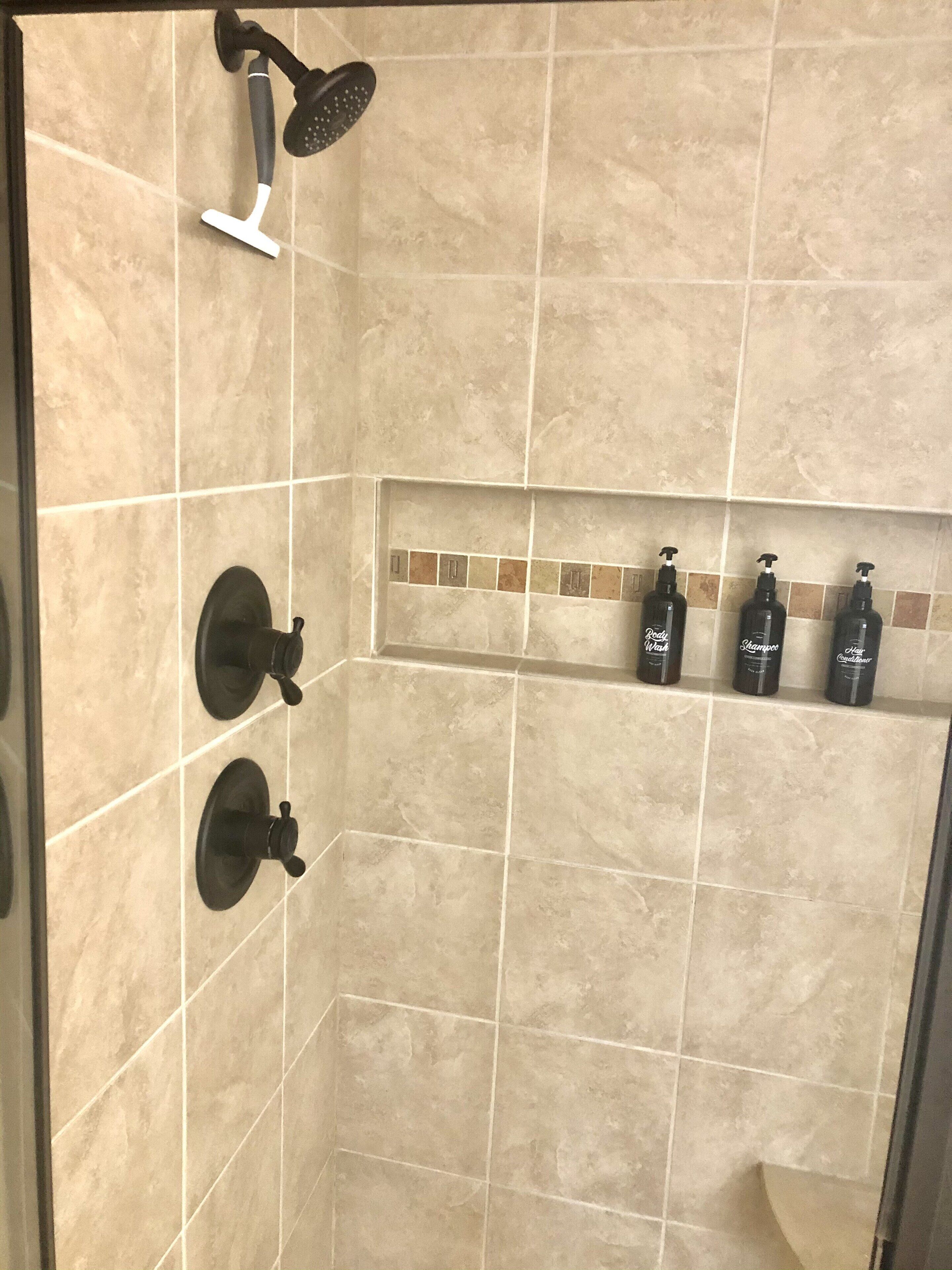 Ein Duschbereich mit beigen Fliesen, einer Handbrause und einem Ablagefach mit drei Shampoo- und Conditionerflaschen.