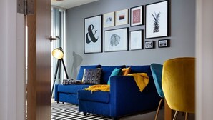 A modern living room with a blue sofa, colorful cushions, and a yellow chair. There are framed artworks on the wall and a floor lamp. - Plymouth Sea-View PENTHOUSE-Habita Property (England)