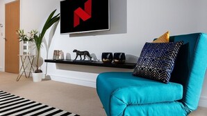 A teal sofa with patterned pillows, a mounted TV with a red logo, a small shelf with decorative items, and a potted plant. - Plymouth Sea-View PENTHOUSE-Habita Property (England)