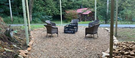 Un espace extérieur aménagé avec des chaises en osier et un barbecue noir, entouré d’un chemin de gravier et de poteaux en bois.