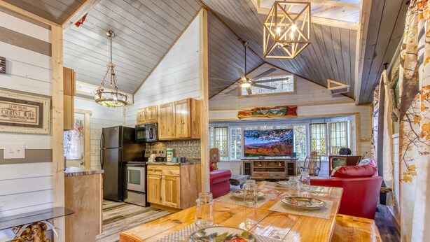 A cozy cabin interior with a wooden dining table set for four, a kitchen with wooden cabinets, and a large window overlooking a scenic landscape.
