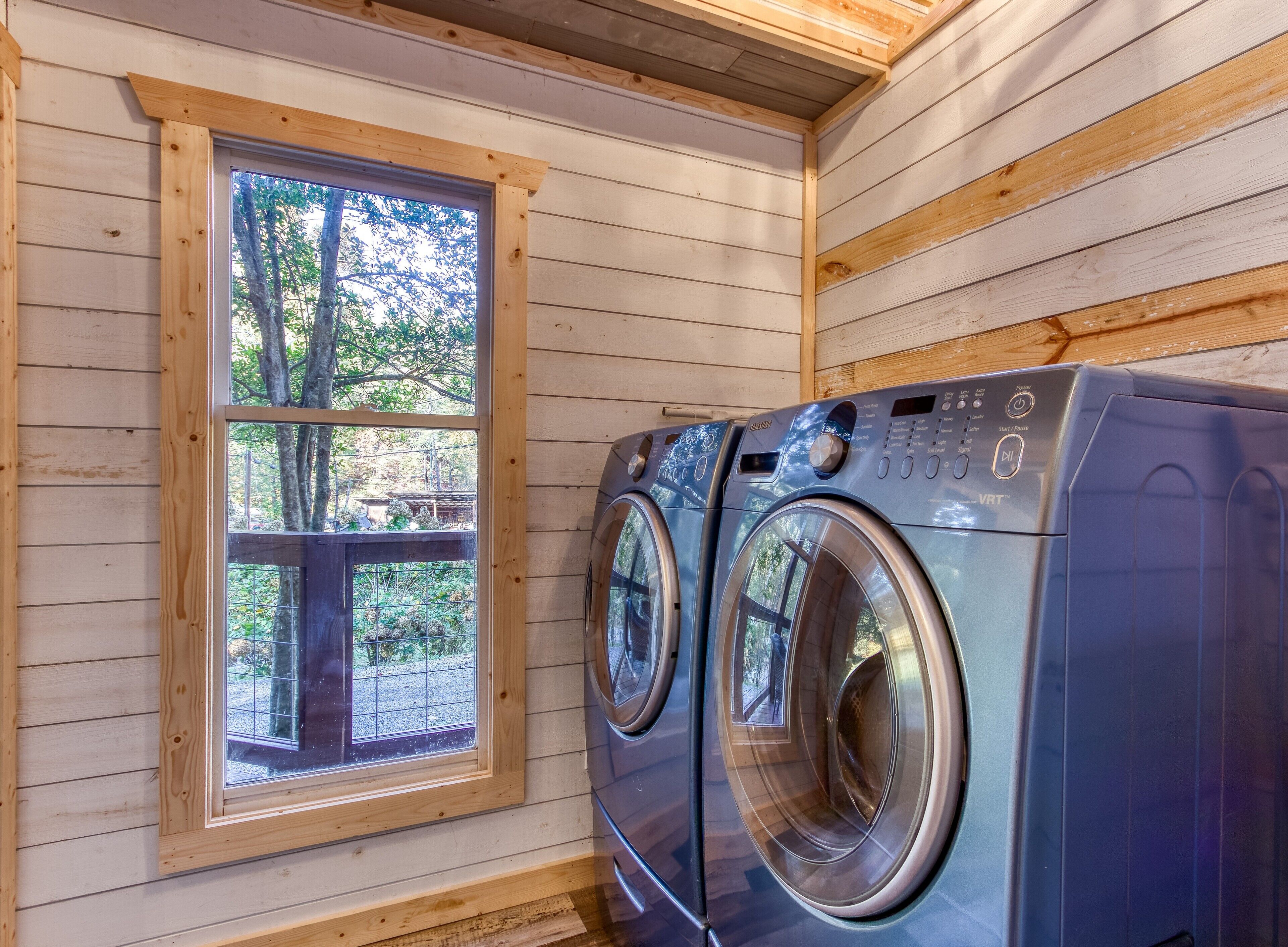A laundry room with two front-loading washing machines, a wooden window, and a view of the outdoors.