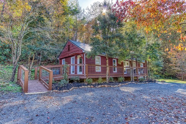 A wooden cabin with a covered porch and a gravel driveway, surrounded by autumn foliage.