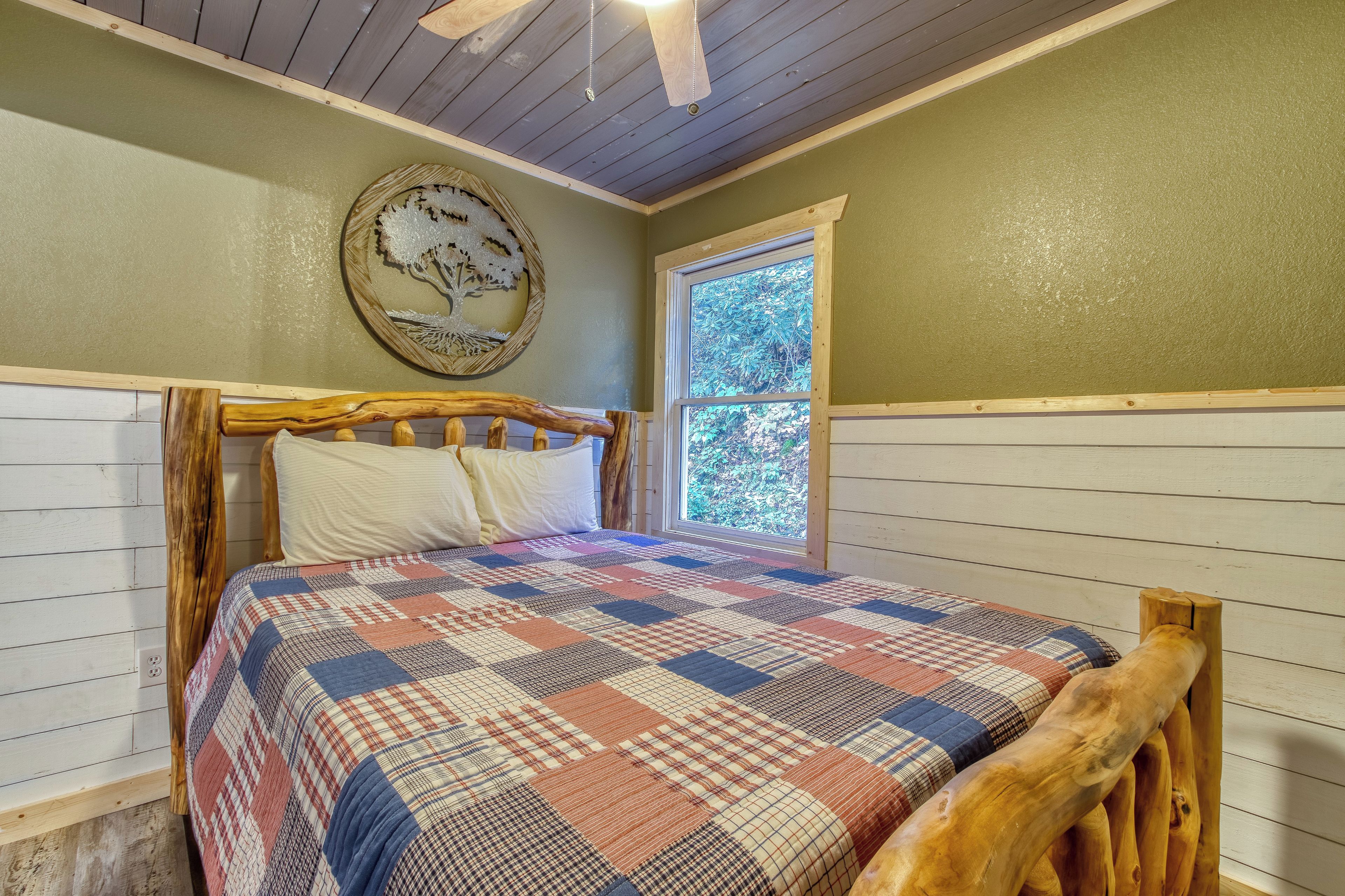 A bedroom with a wooden headboard, a quilted bedspread, a wall clock, and a window with a view of trees.