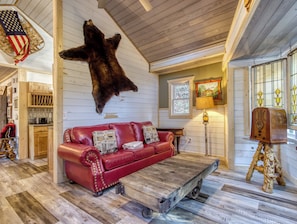 A cozy interior with a red leather sofa, a mounted bear hide, wooden flooring, and a rustic wooden coffee table. - Fire-Pit | Hot Tub | WiFi | Pet Friendly | Arcade | Close to Attractions (Sevierville)
