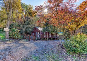 A rustic cabin with a wooden deck and a gravel path leading to it, surrounded by autumn foliage.