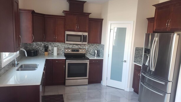 A kitchen with dark wooden cabinets, stainless steel appliances, a tiled backsplash, and a glass door leading to a hallway. - Stylish Heritage Apt Ideal For Long Stays - 3 BR with Jacuzzi (Fort McMurray)