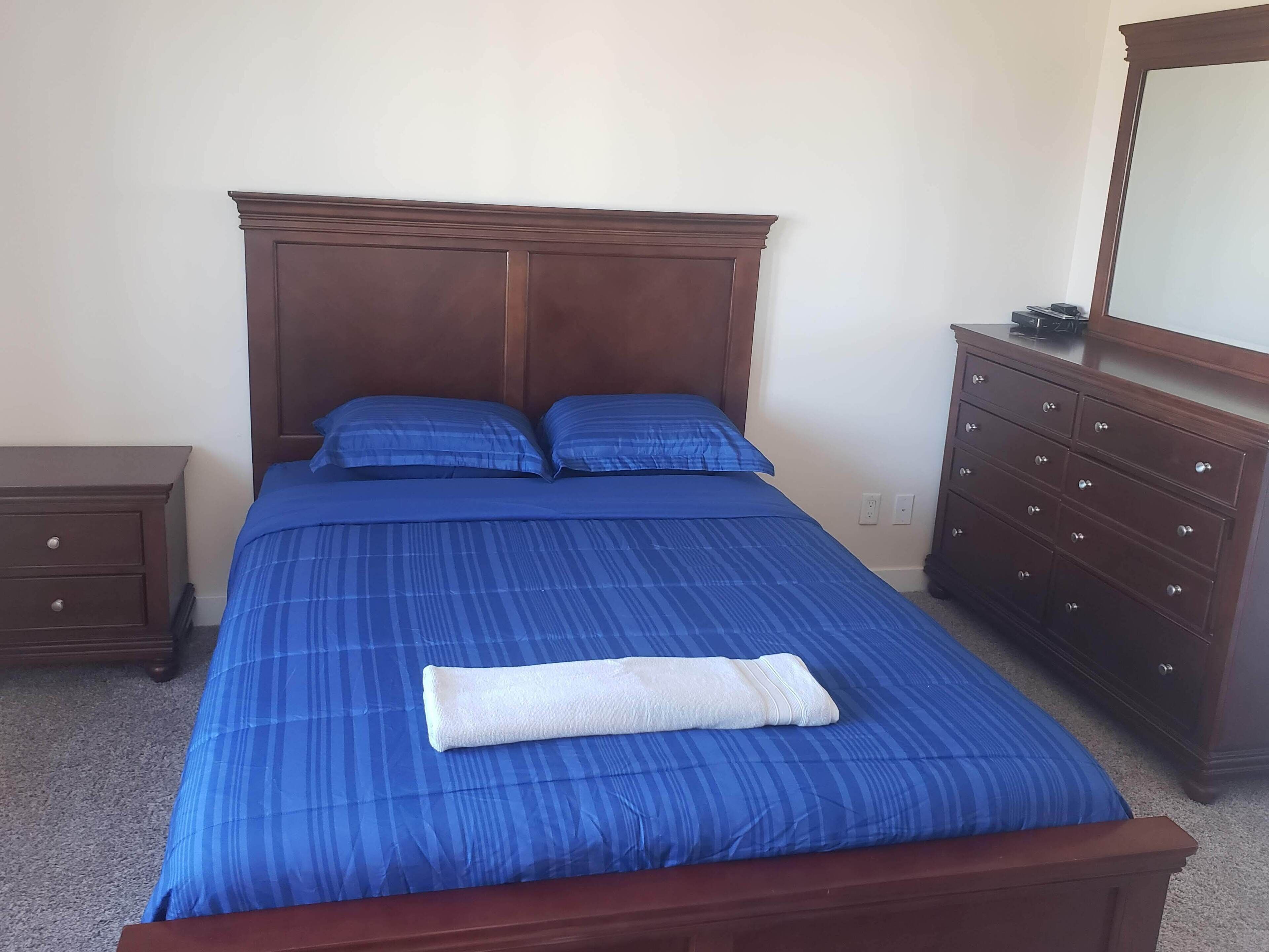 A neatly made bed with blue bedding and a white towel, a wooden nightstand with drawers, a dresser with multiple drawers, and a mirror on the wall.