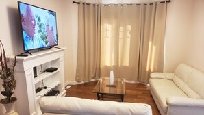 A living room with a white sofa, a glass coffee table, a flat-screen TV on a white stand, and a potted plant. - Stylish Heritage Apt Ideal For Long Stays - 3 BR with Jacuzzi (Fort McMurray)