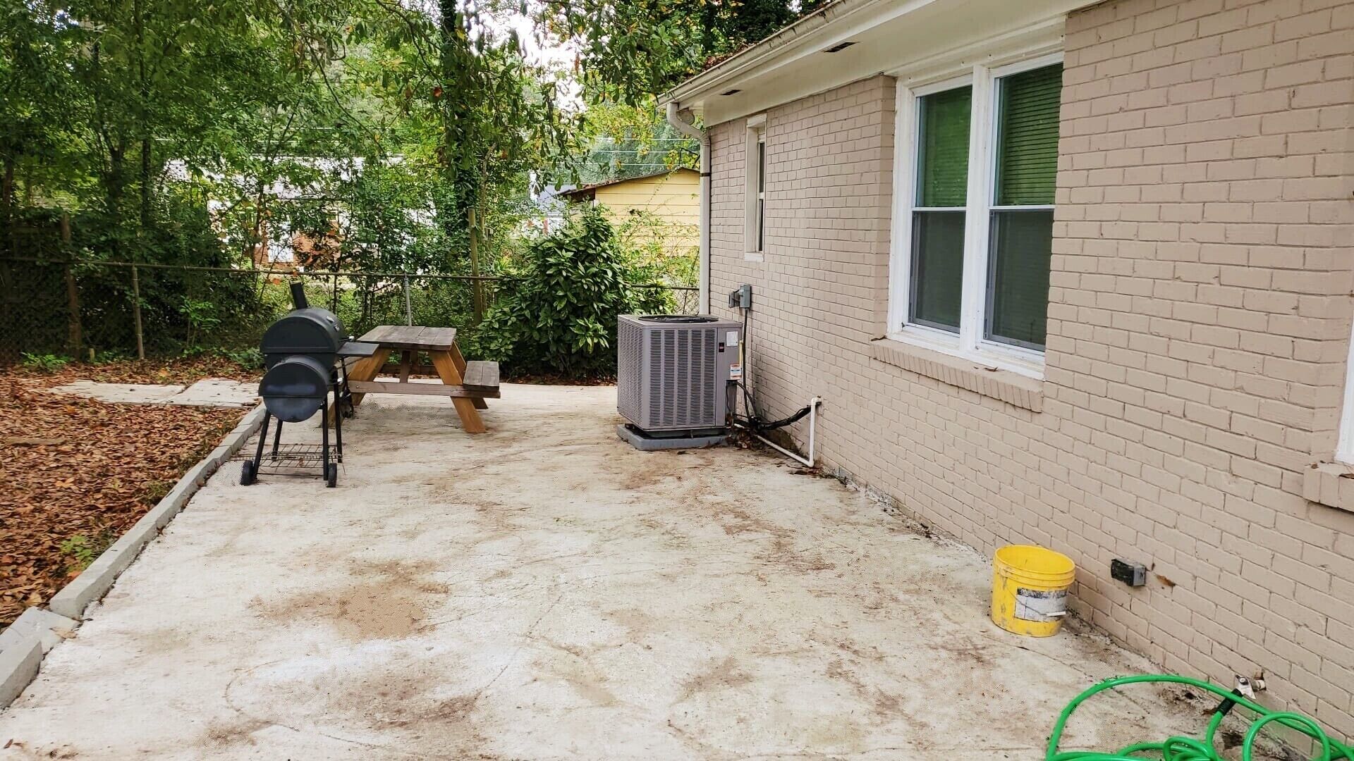 A backyard with a barbecue grill, a picnic table, an air conditioning unit, and a yellow bucket.