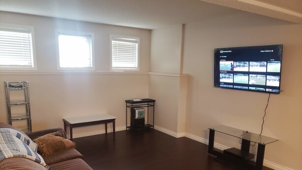 A living room with a brown sofa, a glass coffee table, a TV mounted on the wall displaying multiple channels, and a window with blinds. - 3 Bedroom Furnished Apartment  suitable for Company Staff with Large Yard  Space (Fort McMurray)