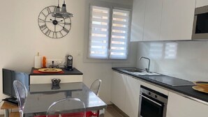A modern kitchen with a dining area, including a table with chairs, a clock on the wall, and a window with blinds.