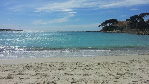 A beach with clear turquoise water, a sandy shore, and a building with trees on a cliff.