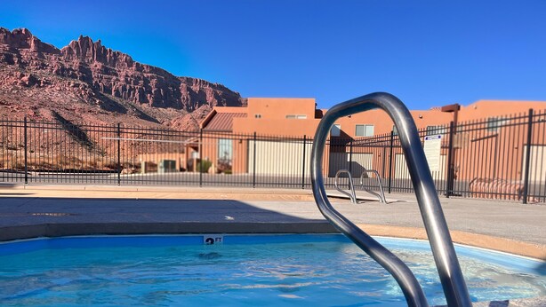 A swimming pool with a metal ladder, a desert landscape, and a building in the background. - 18A4 Moab Adventure! Pool with Red Rock Views (MOAB)
