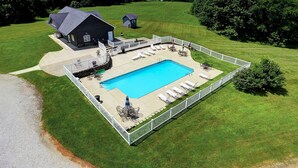 Una gran piscina rectangular con solárium, rodeada por una valla blanca. Hay sillas de descanso y una pequeña zona de patio con mesas y sillas.