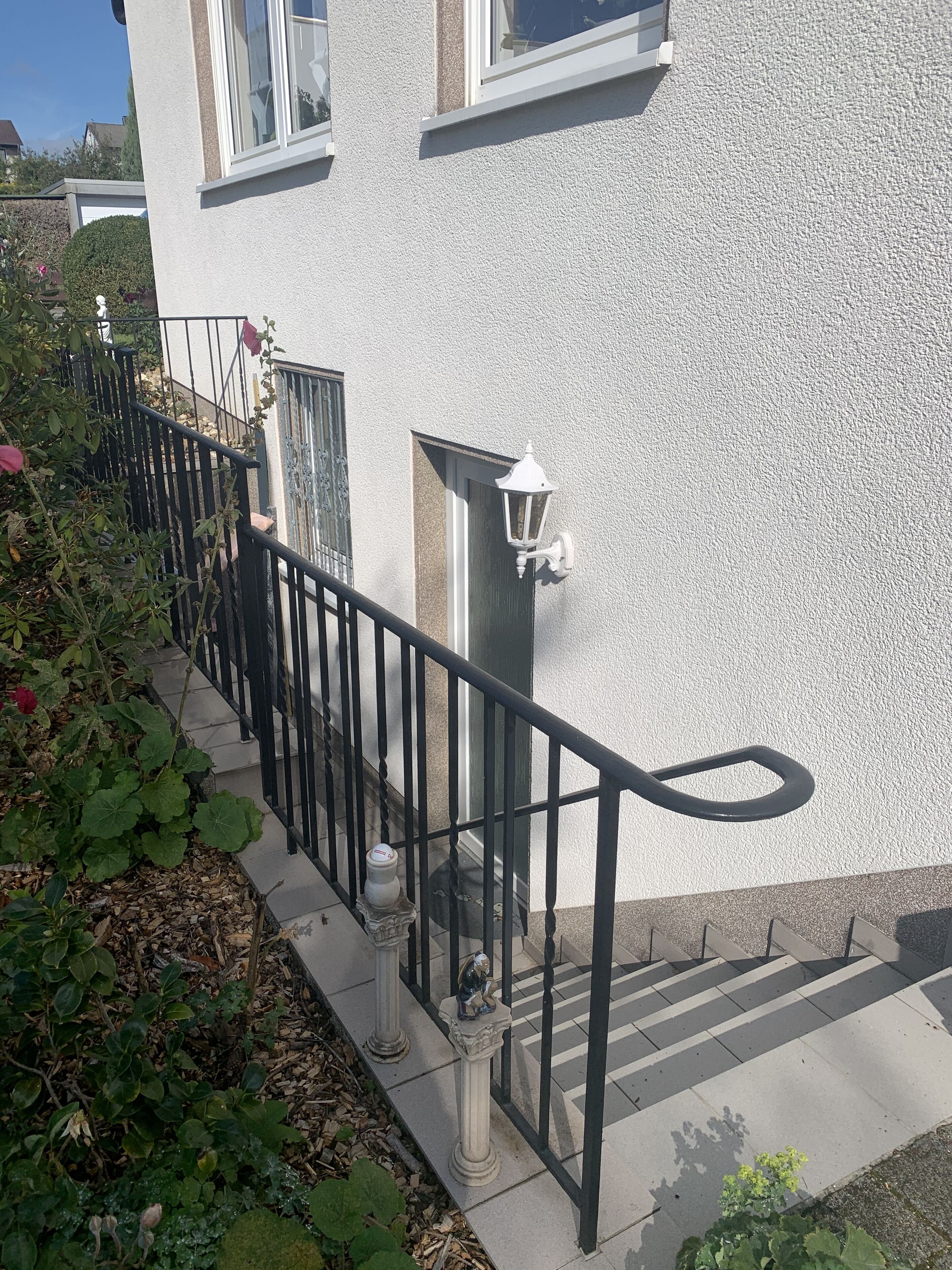 A residential building with a white exterior, a black metal railing, and a small garden area with plants and a red flower.