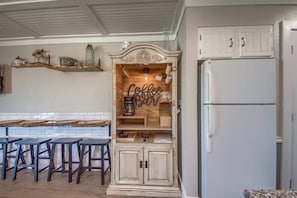 A kitchen with a white refrigerator, wooden bar stools, a coffee bar, and decorative items on shelves.
