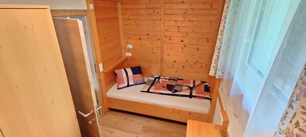 A small wooden room with a bunk bed, a door, and a window with floral curtains. - Wohnung Ost - Haus Lukasser (Kosten)