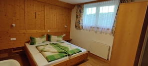 A bedroom with a wooden headboard, a bed with green and white bedding, a window with sheer curtains, and a radiator.