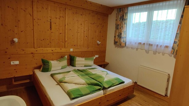 Une chambre à coucher avec une tête de lit en bois, un lit avec une literie verte et blanche, une fenêtre avec des rideaux légers et un radiateur.
