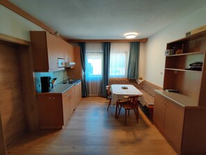A compact kitchen with wooden cabinets, a dining table, and a window with curtains. - Wohnung Ost - Haus Lukasser (Kosten)