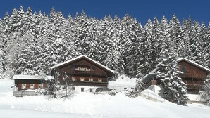 Un chalet enneigé, composé de plusieurs cabanes en bois, entouré d’une forêt dense de conifères.