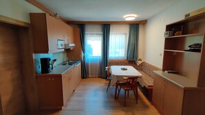 A compact kitchen with wooden cabinets, a dining table, and a window with curtains. - Wohnung Ost - Haus Lukasser (Kosten)