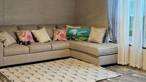 A beige sectional sofa with patterned cushions and a matching throw, positioned on a patterned rug in a room with a large window and curtains. - MANATEE WATCH DEN 1 BEDROOM PET FRIENDLY (Crystal River)
