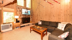 A cozy living room with a stone fireplace, a television mounted above it, a sofa with a 'SKI LODGE' pillow, and skis hanging on the wall. - Spacious Kearsarge Woods Unit w Association Pool & Tennis 2 Mins to Downtown (North Conway)