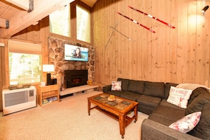 Un salon chaleureux avec une cheminée en pierre, un téléviseur fixé au-dessus, un canapé agrémenté d’un coussin orné de l’inscription « SKI LODGE », et des paires de skis accrochées au mur.