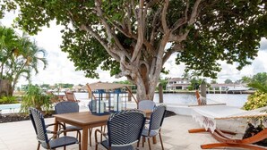 An outdoor patio with a wooden table and chairs, a hammock, and a large tree providing shade. - Beautiful Waterfront Condo on Canal with Pool & Walk to Beach! Boat Watch! 2b/2b (Pompano Beach)