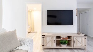 A living room with a flat-screen TV mounted on the wall, a light-colored sofa, and a wooden entertainment unit with a plant. - Beautiful Waterfront Condo on Canal with Pool & Walk to Beach! Boat Watch! 2b/2b (Pompano Beach)